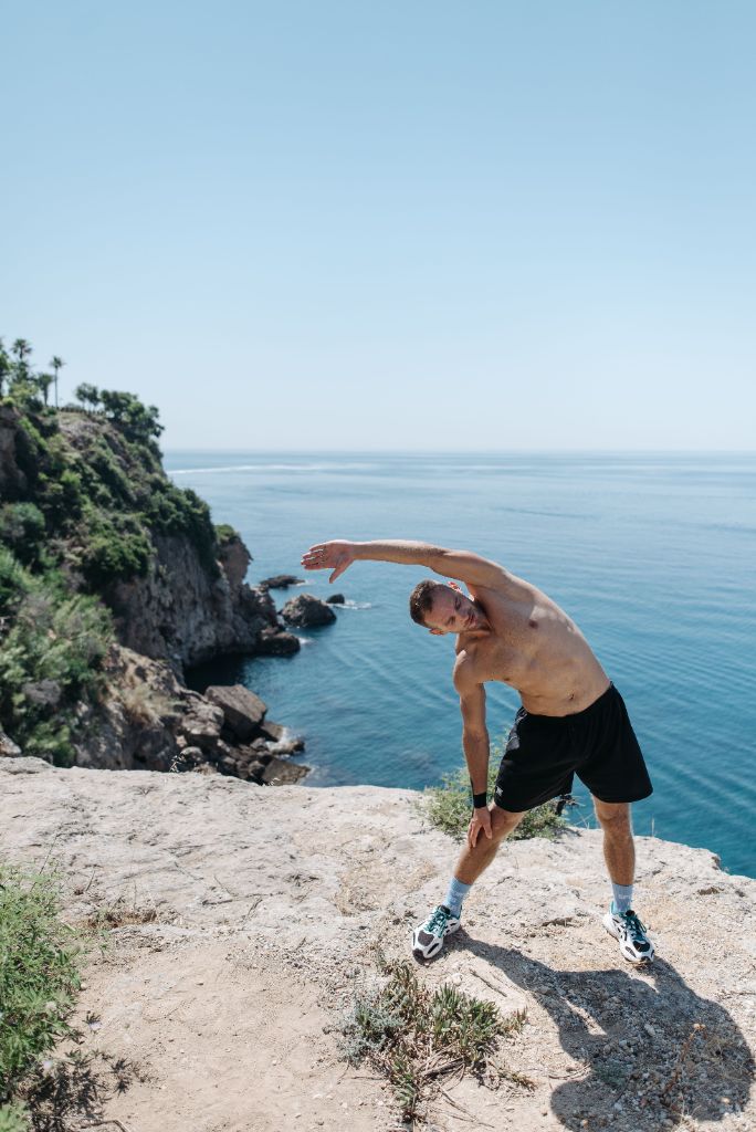 Shirtless man stretching on a sun-bright ocean cliff with turquoise water below